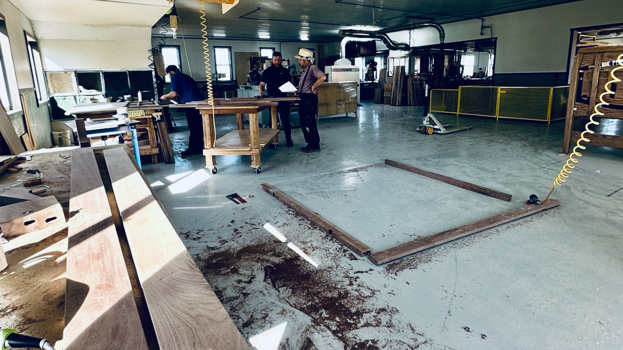 Men in a factory workshop, one holding a paper, surrounded by wooden planks and a wooden table with wheels on a concrete floor.