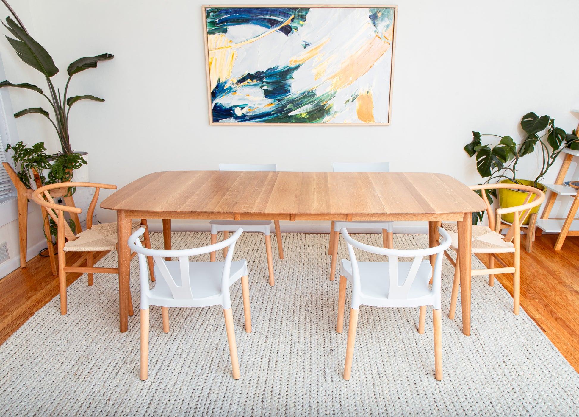 The Mar Vista Scandinavian extendable Oval Dining Table shown with white chairs in a room, surrounded by plants and a painting on the wall.